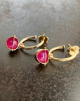 A pair of gold hoop earrings with pink ruby cabochon gemstone hoop charms, displayed on a dark textured surface.