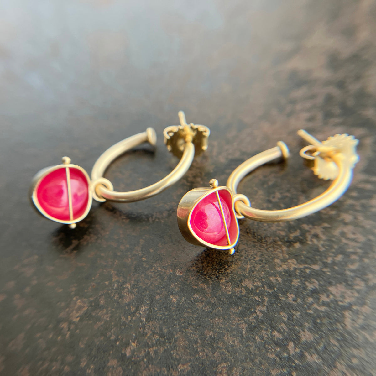 A pair of gold huggie hoop earrings with a small, round pink ruby gemstone hoop charms resting on a dark textured surface.