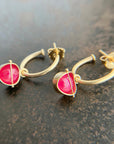 A pair of gold huggie hoop earrings with a small, round pink ruby gemstone hoop charms resting on a dark textured surface.