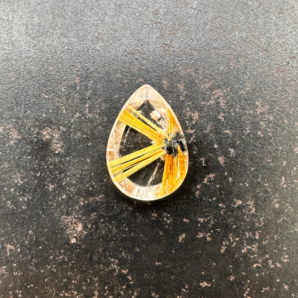 A teardrop-shaped gemstone featuring a striking golden-yellow radial pattern with a small dark inclusions, centered on a dark surface.
