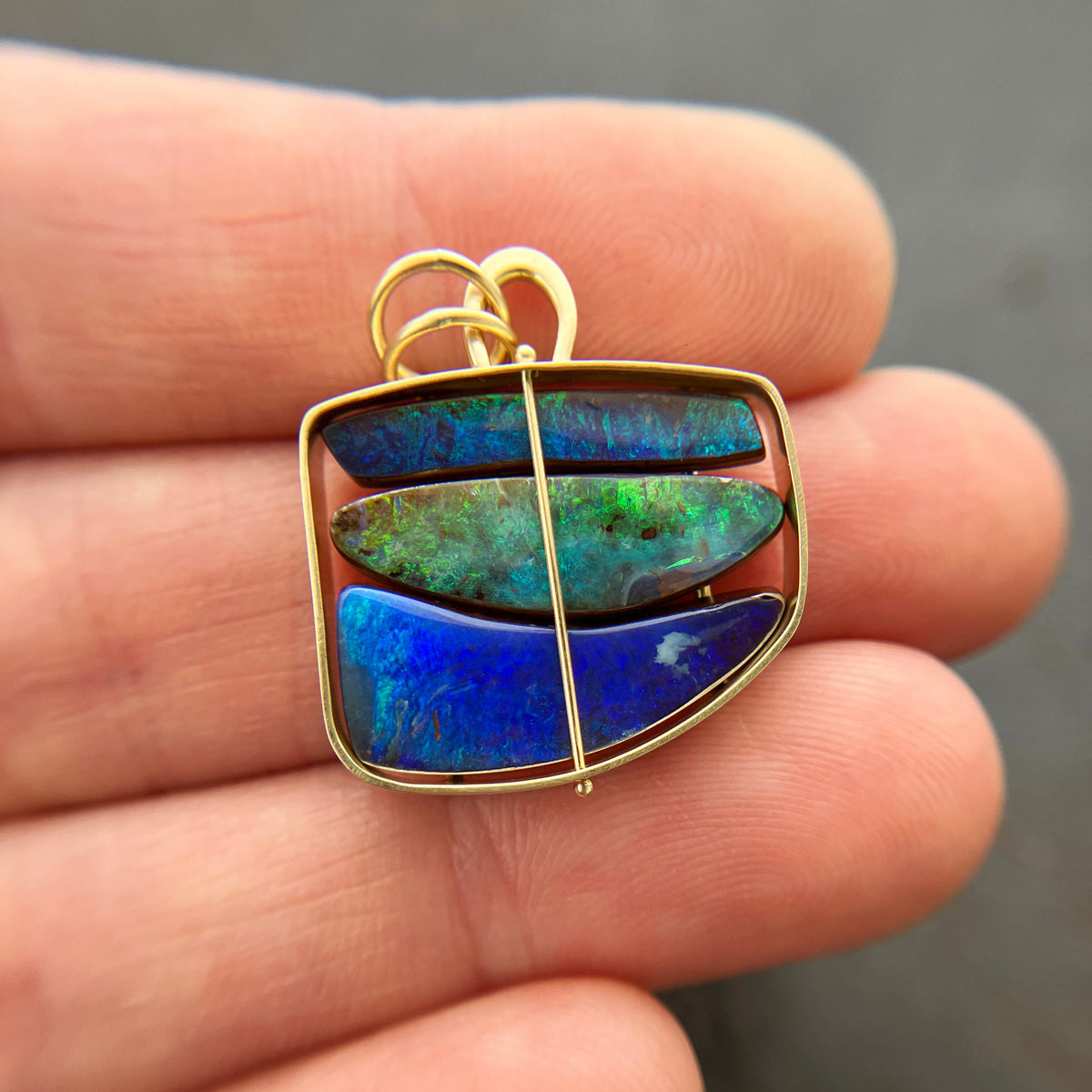 A gold pendant with three horizontal iridescent blue-green opal inlays, held in the palm of a hand.