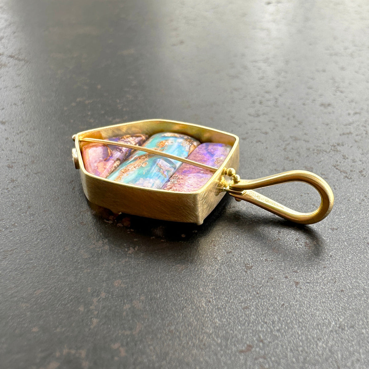 An 18k gold pendant with a loop bale, featuring three wood replacement opals showing purple, blue, and green hues, resting on a dark gray textured surface.