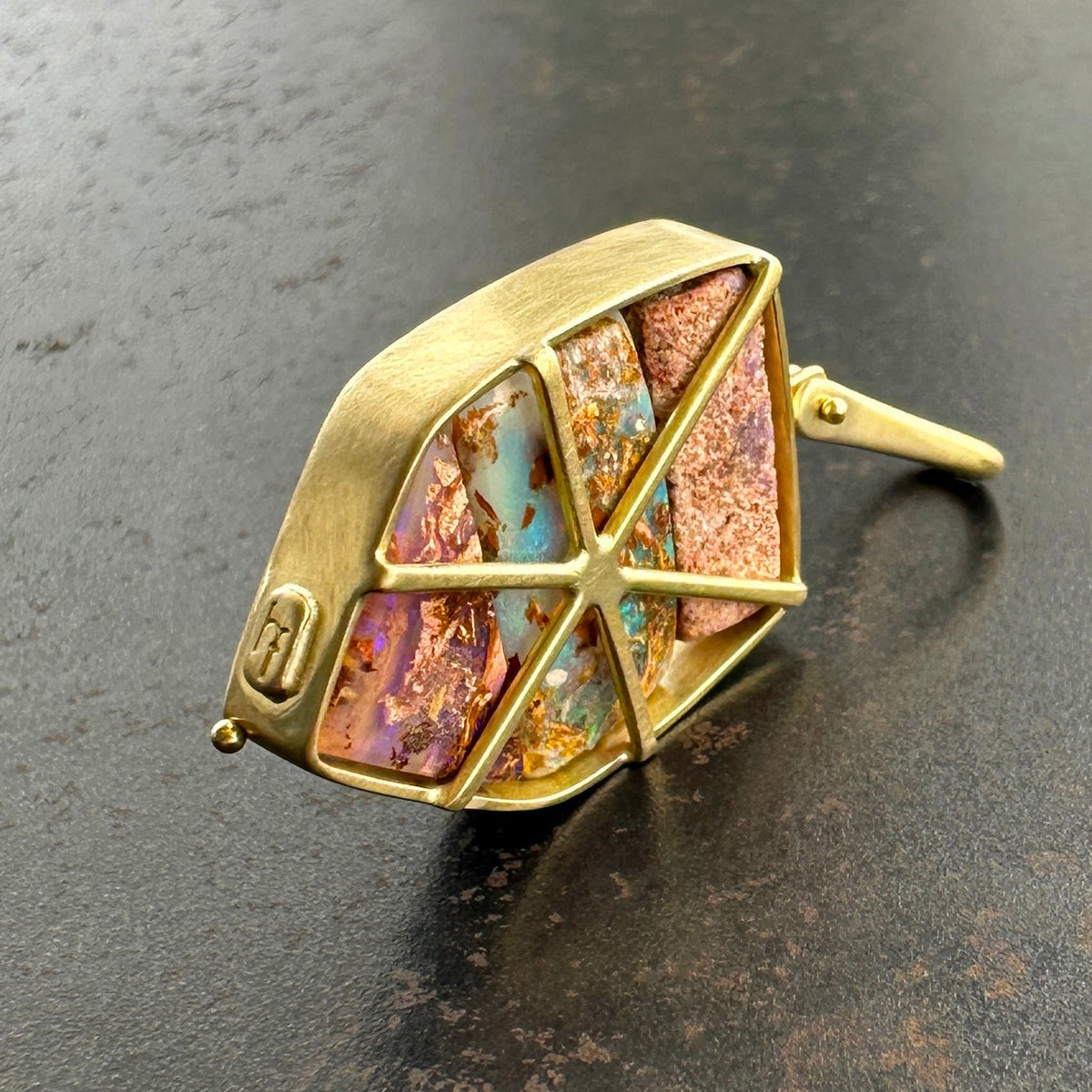 An 18k gold geometric pendant with wood replacement opals in shades of pink, blue, and purple, displayed on a dark textured surface.