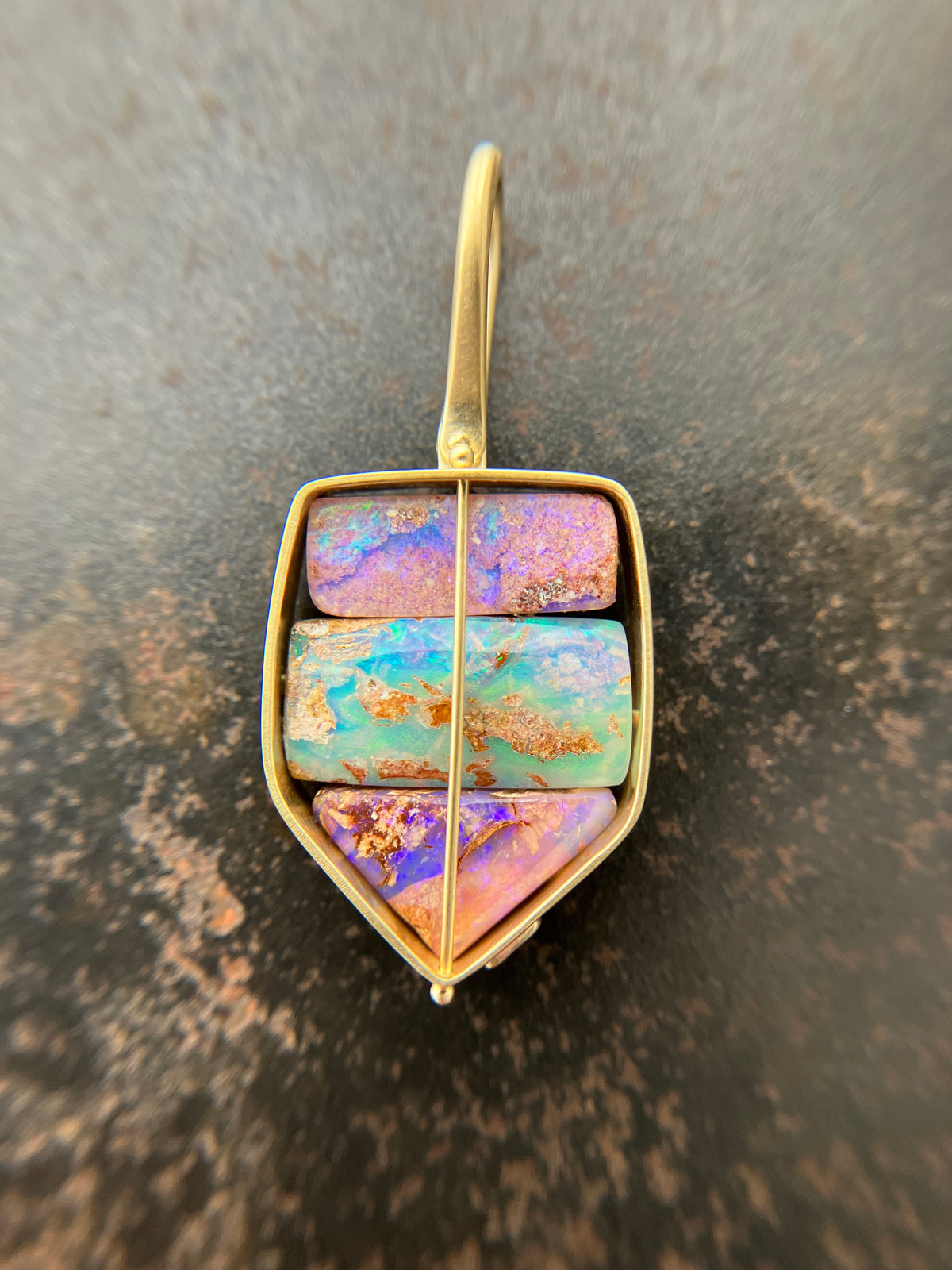 A shield-shaped pendant featuring an 18k gold frame and three rectangular inlay wood replacement opals in pink, turquoise, purple, and brown.