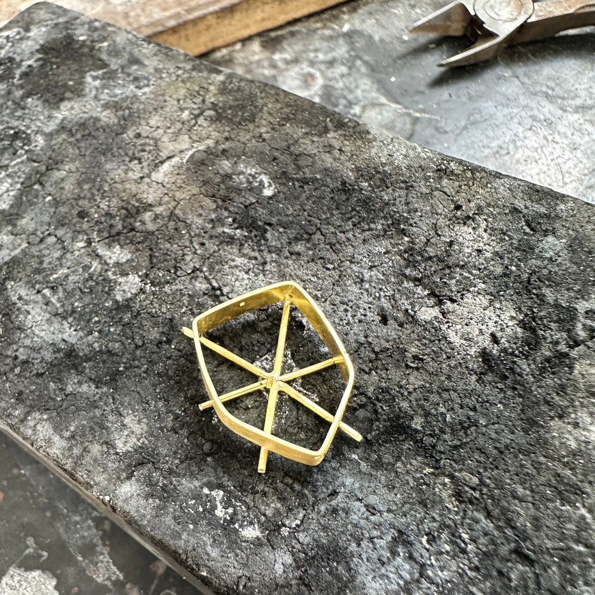 A small gold metal jewelry component or frame piece resting on a dark, textured stone surface with metal tools visible in the background.