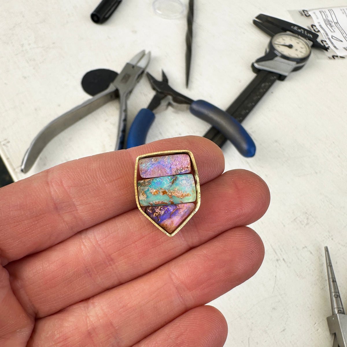 A shield-shaped pendant featuring an 18k gold frame and three rectangular inlay wood replacement opals in pink, turquoise, purple, and brown.