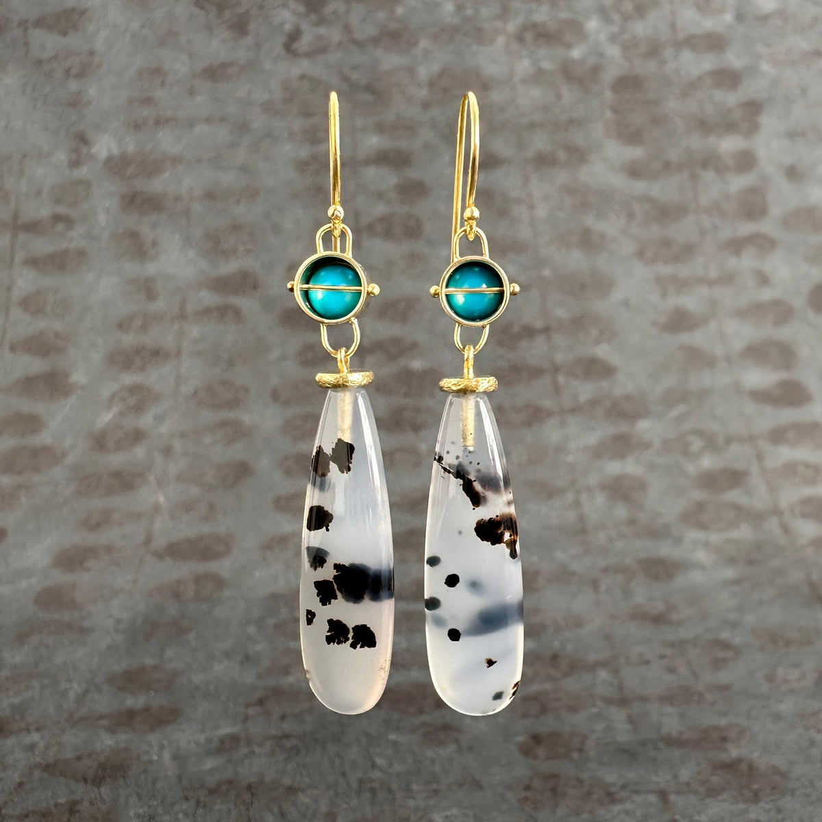 Pair of earrings with teardrop-shaped montana agates and turquoise with 18k gold on a textured gray background