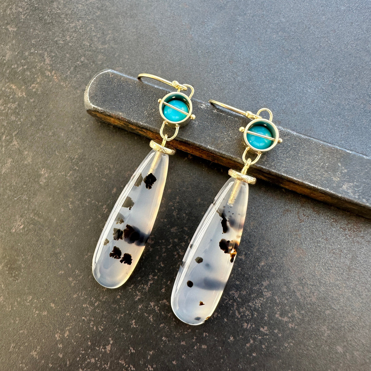 Pair of earrings with teardrop-shaped montana agates and turquoise with 18k gold on a textured gray background