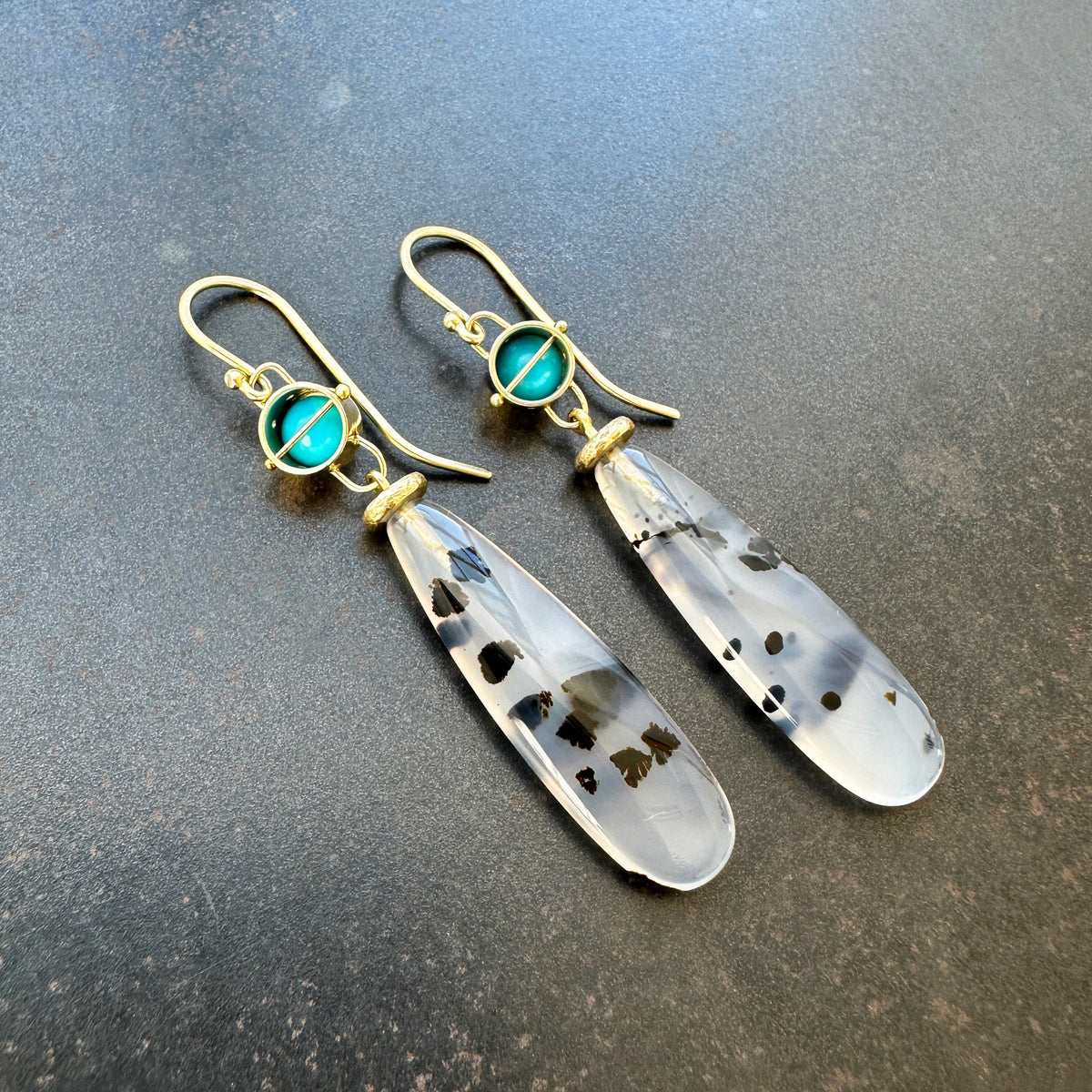 Pair of earrings with teardrop-shaped montana agates and turquoise with 18k gold on a textured gray background